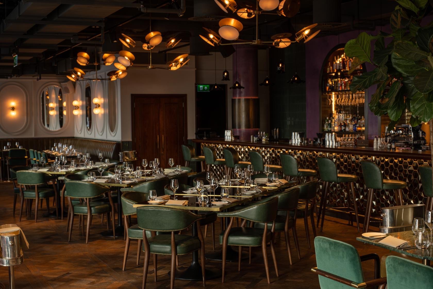 A rooftop restaurant with green velvet chairs, set dining tables, and a backlit bar at Wagtail.