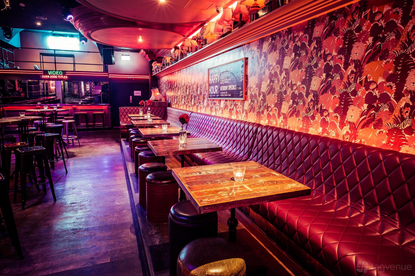 An event space in a bar with tufted red leather banquette seating, wooden tables, and floral wallpaper at Simmons | Oxford Street.