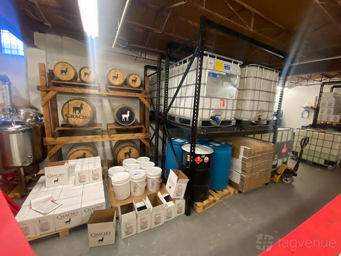A distillery bar with wooden barrels, stacked blue drums, and metal shelving at Chacho Spirits.