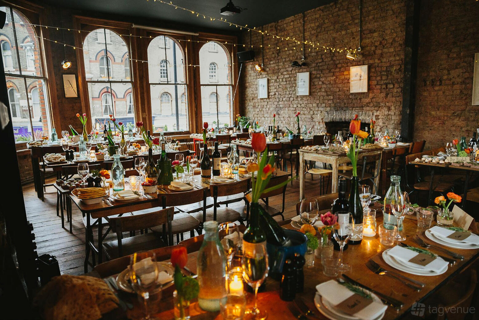A bar with exposed brick walls, large arched windows, wooden tables set with candles and glassware at The Elgin.