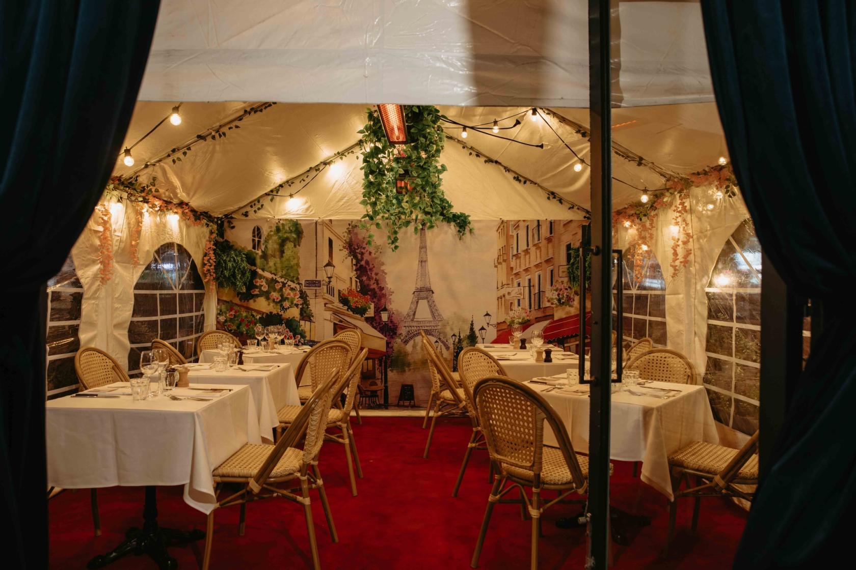 An outdoor dining area with bistro chairs, white tablecloths, string lights, and a Paris-themed mural at Ouest France French Bistro.