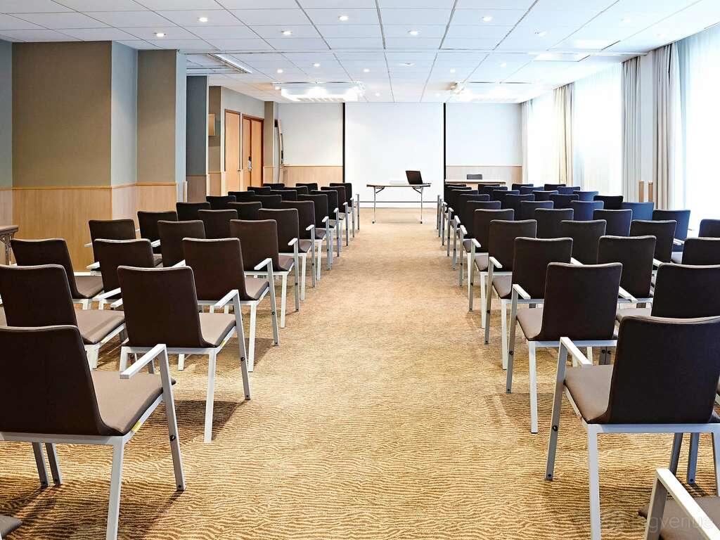 A hotel meeting room with rows of black chairs facing a projection screen and table, carpeted floor, and large windows at Novotel Leeds Centre.