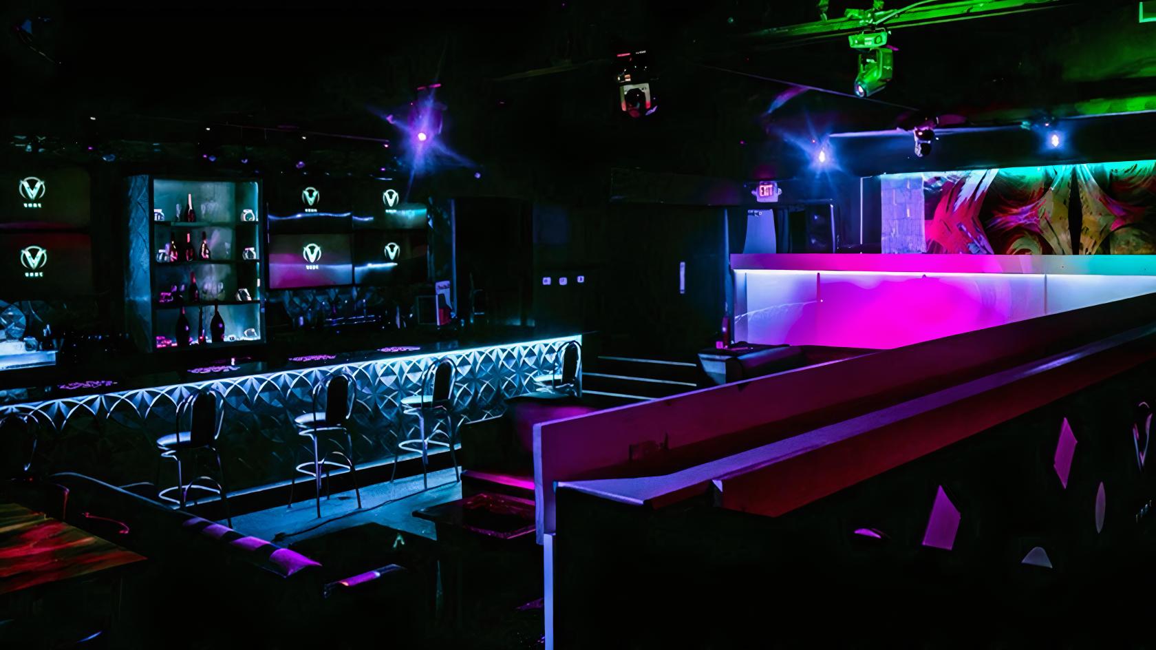 A bar in a restaurant with tufted seating, neon lighting, backlit shelves, and bar stools at VODS Lounge and Bistro.