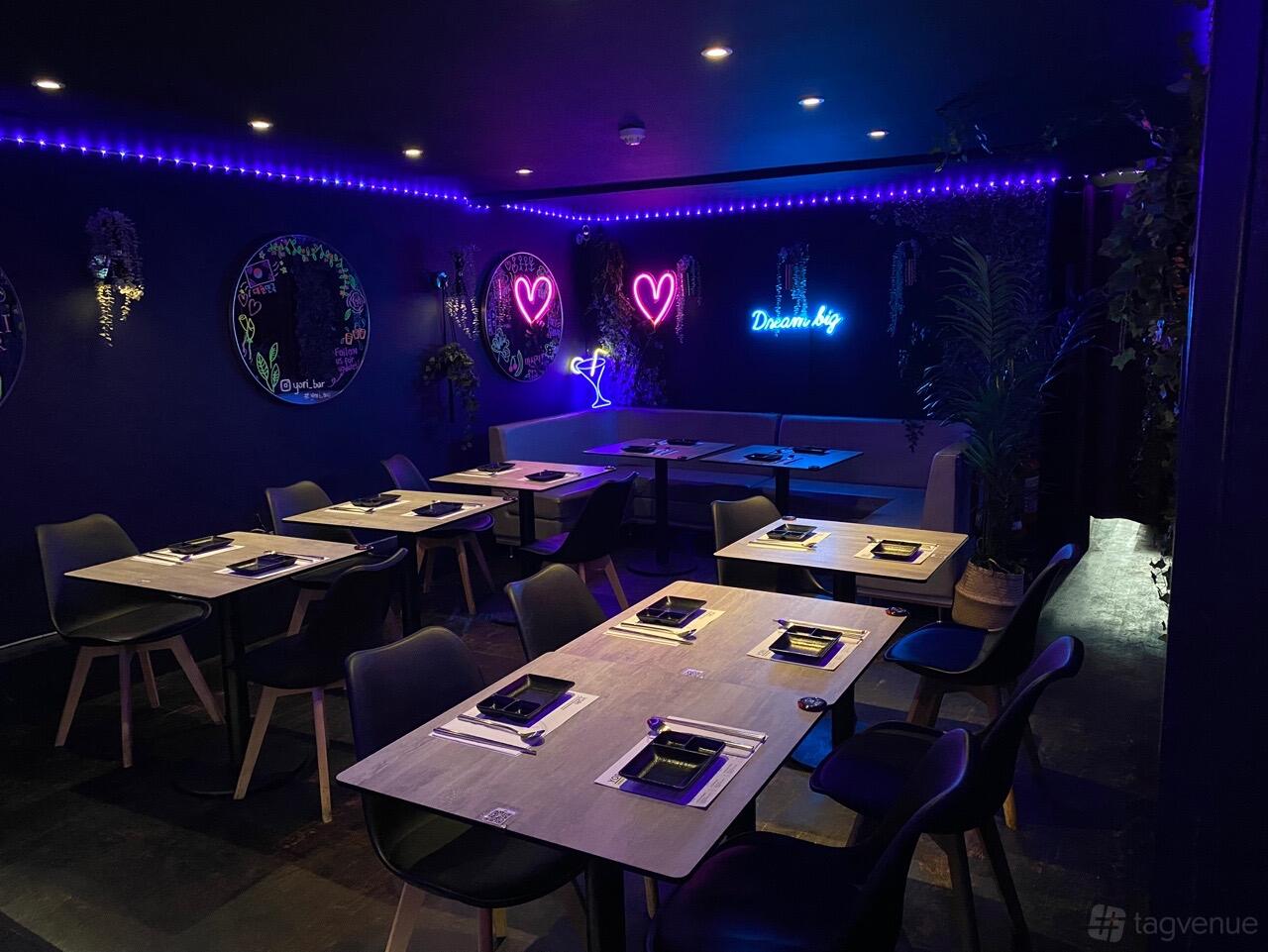 A basement dining area with neon signs, potted plants, and wooden tables set for guests at YORI Korean BBQ - Clapham.