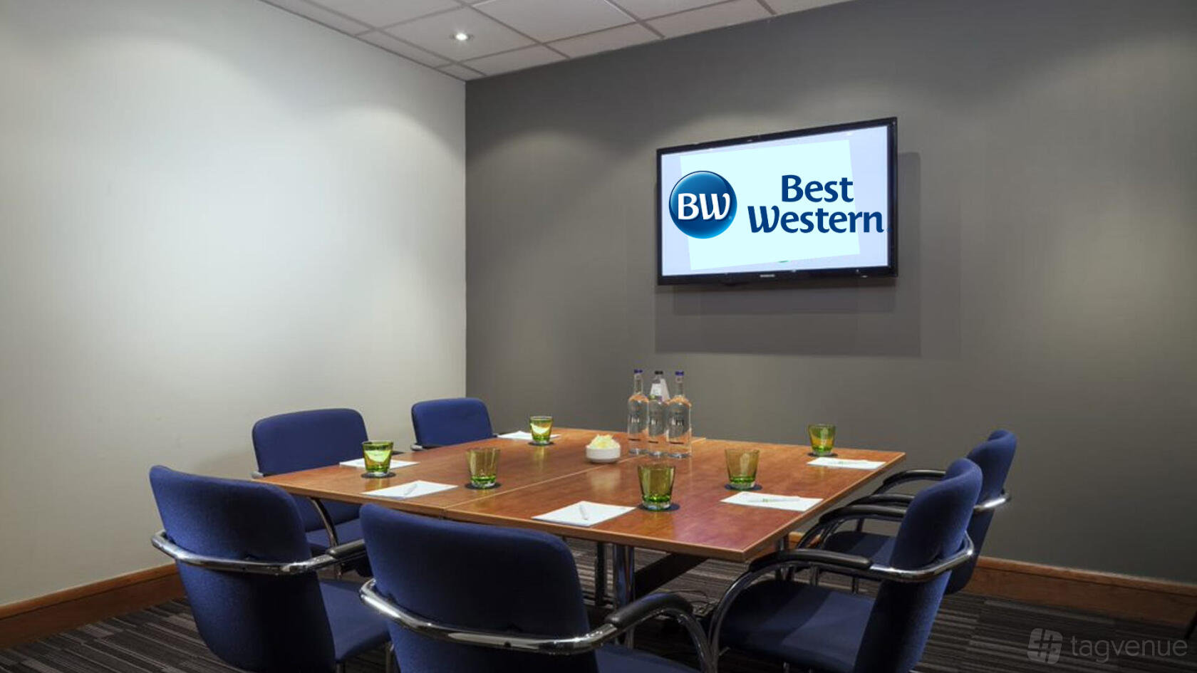A hotel meeting room with a wooden conference table, blue chairs, and a wall-mounted screen at Best Western Ariel London Heathrow.
