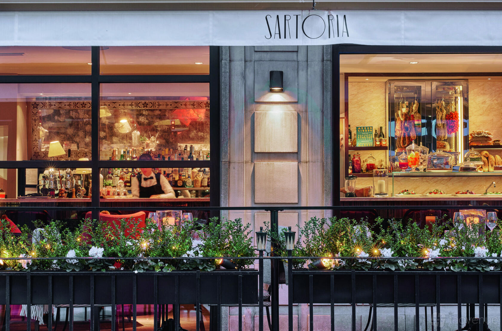 A restaurant exterior with large windows, floral planters, and visible shelves displaying Italian goods at Sartoria.