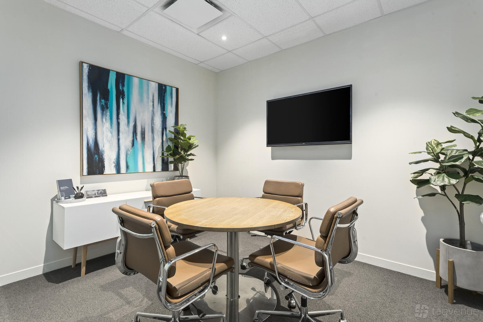 A meeting room with a round wooden table, tan chairs, wall-mounted TV, and abstract artwork at Corporate One.