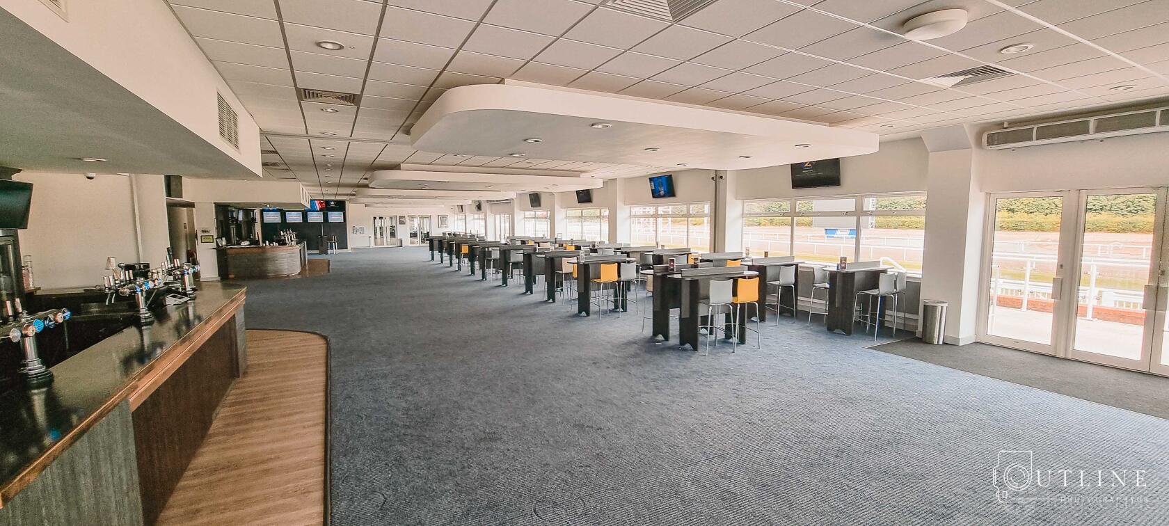 An event venue with high-top tables, bar seating, large windows, and multiple wall-mounted TVs at Chelmsford City Racecourse.