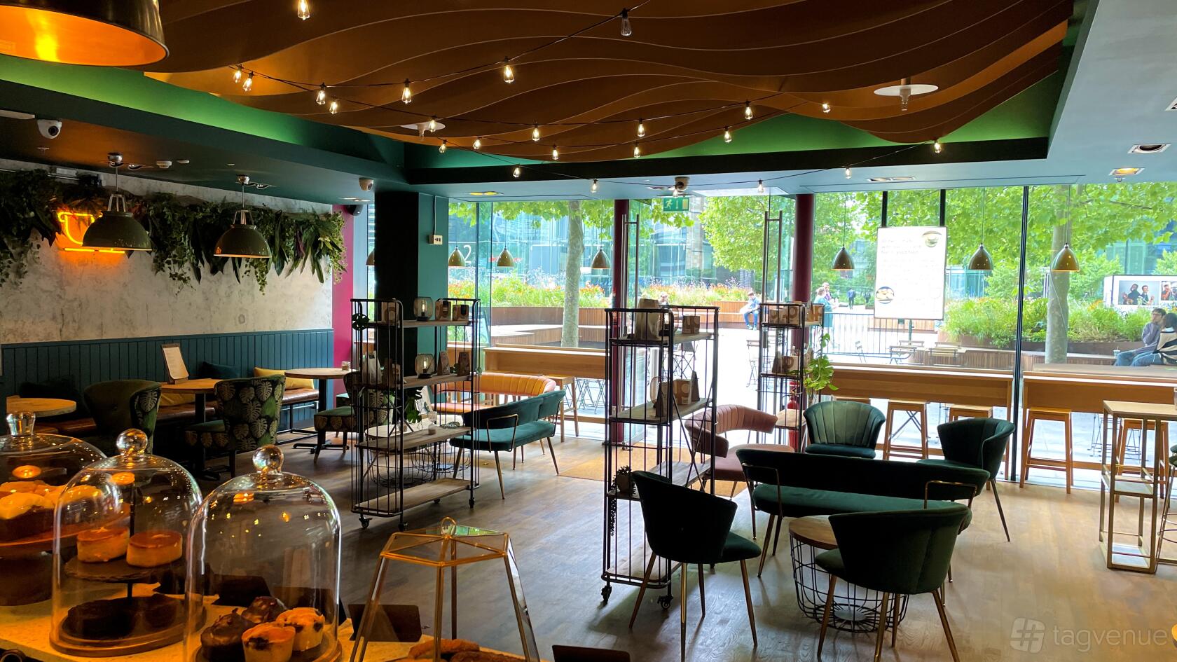 A cafe with floor-to-ceiling windows, indoor plants, string lighting, and assorted lounge seating at BloomsYard - Regents Place.