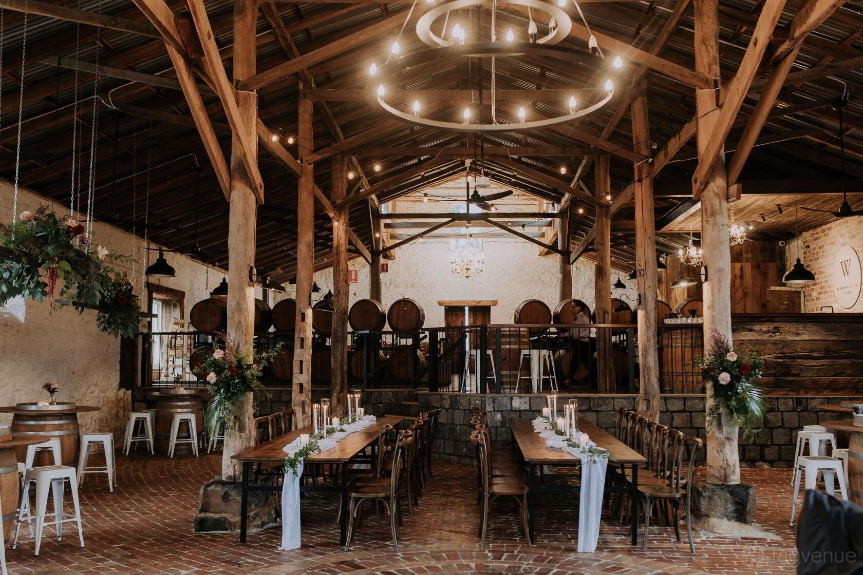 A barn with exposed wooden beams, brick flooring, long wooden tables, and barrel decor at Warrawong Estate.