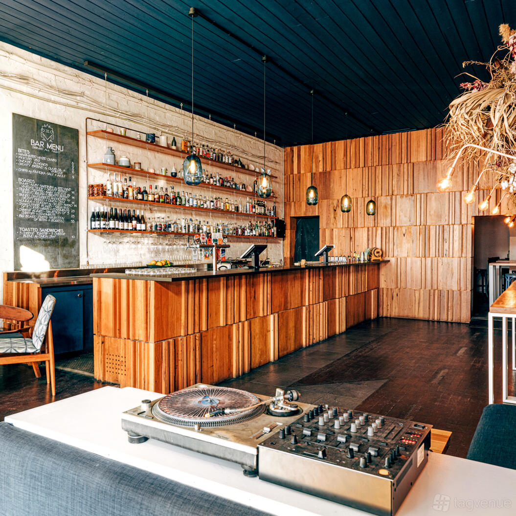 A bar with wooden paneling, open shelves displaying bottles, pendant lights, and a DJ turntable at Bowerbird Bar.