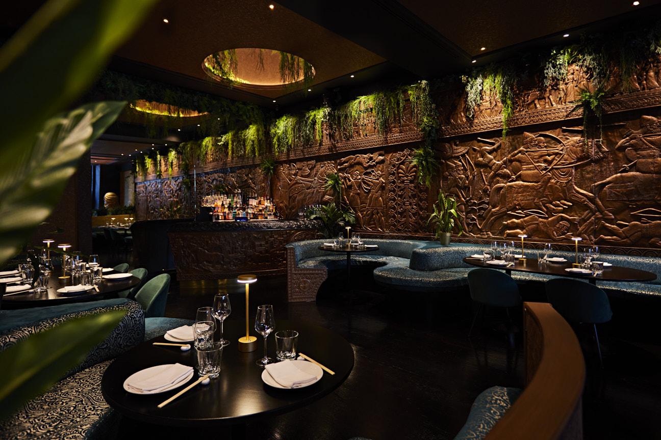 A restaurant interior with carved wall panels, lush greenery, curved banquette seating, and set tables at Gilgamesh London Covent Garden.