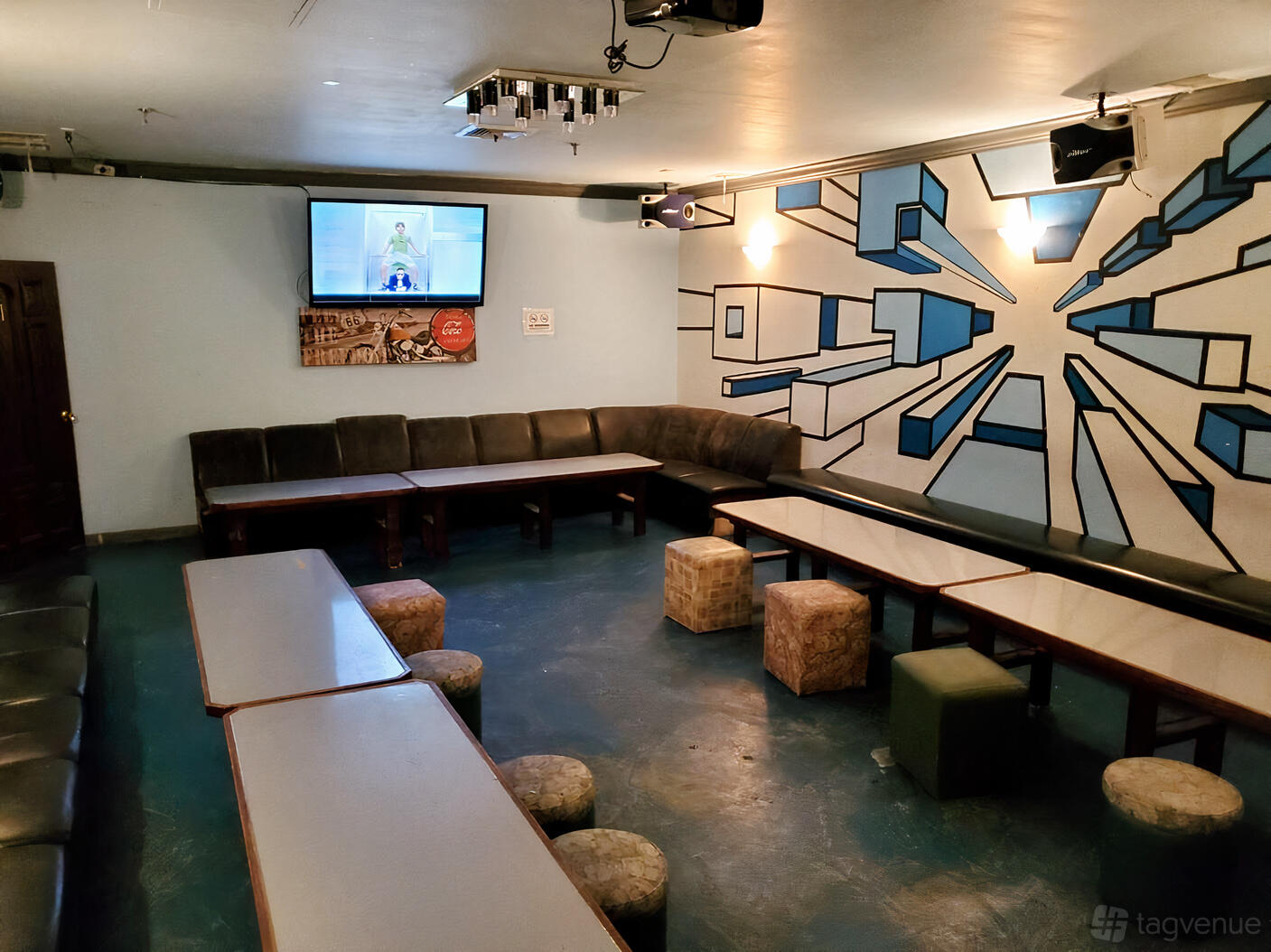 A karaoke bar room with corner sofas, low tables, cube seats, a wall mural, and a TV screen at Christmas Karaoke.