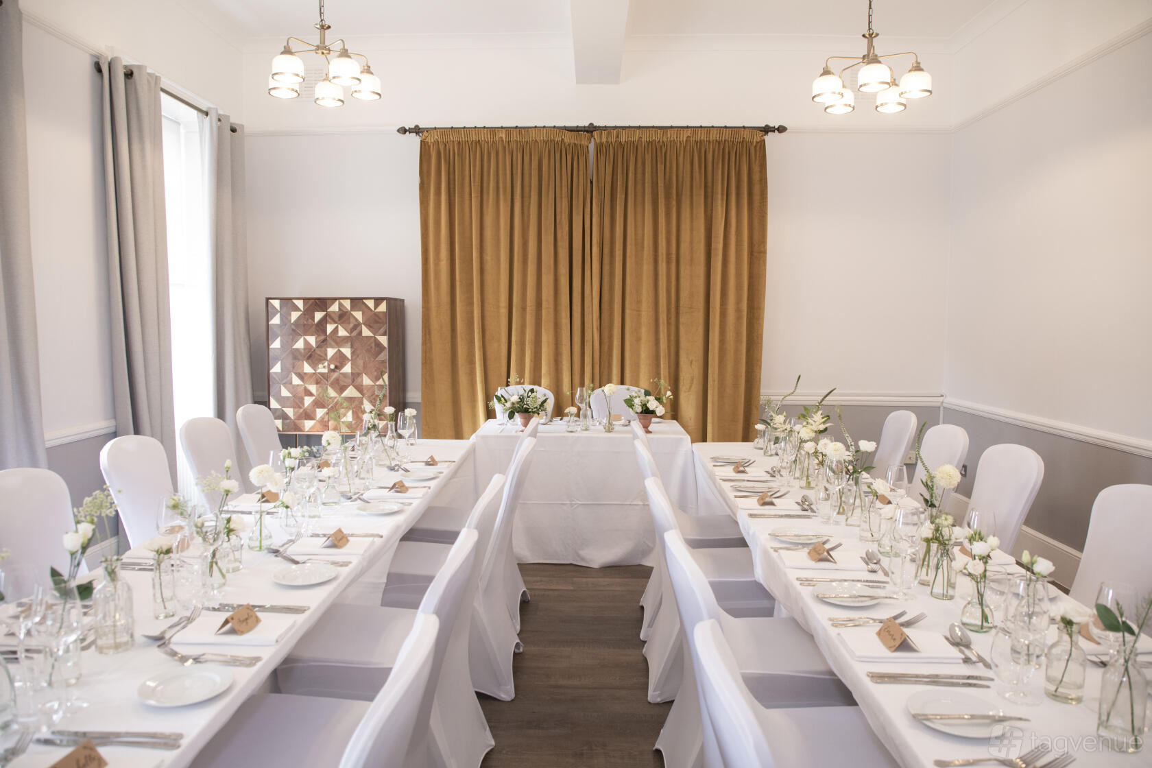 A function room with banquet tables dressed in white linens, floral centerpieces, and gold curtains at Linden House Riverside.