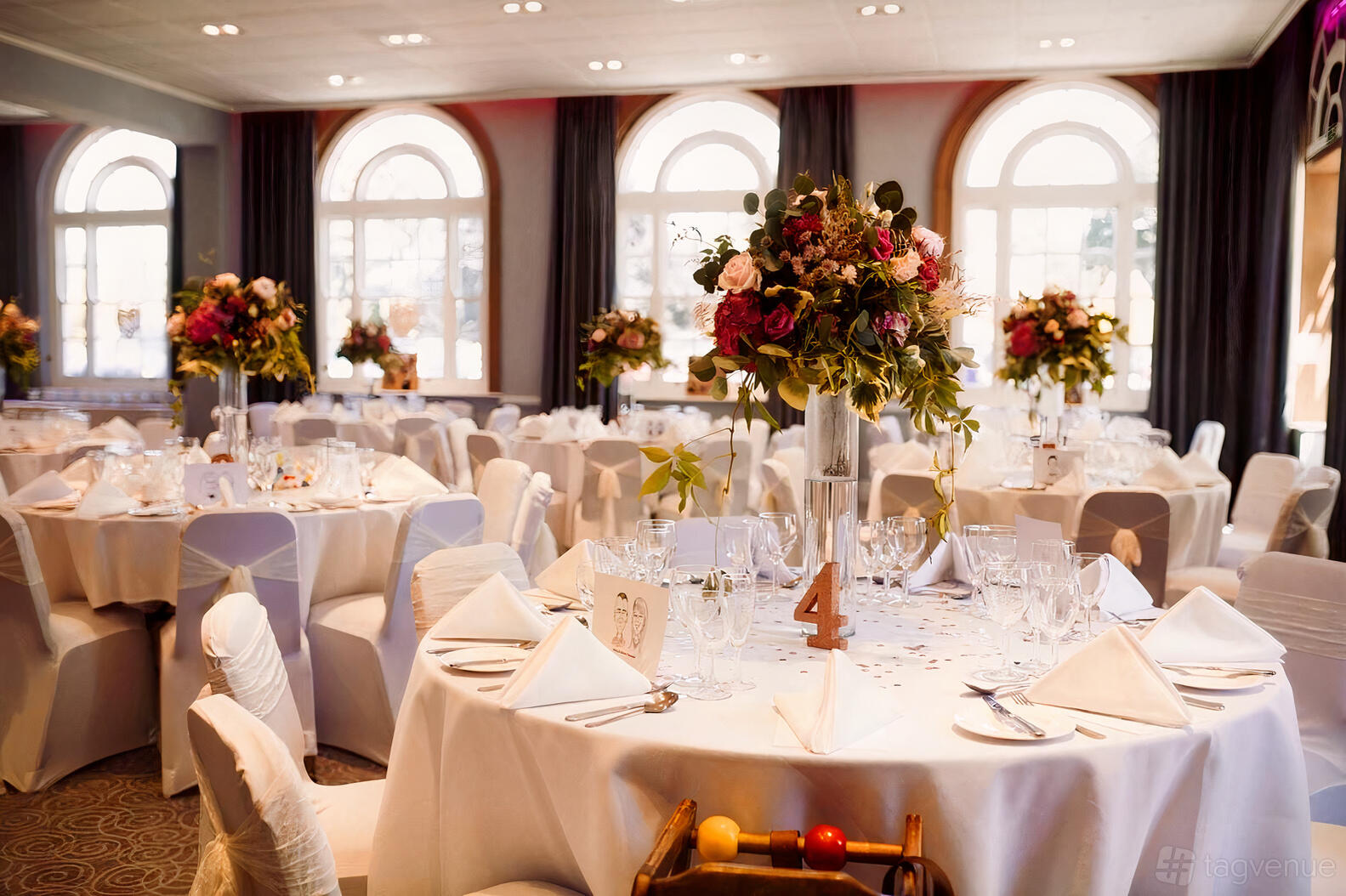 An event venue with round tables, white linens, floral centerpieces, and arched windows at Hawkstone Park.