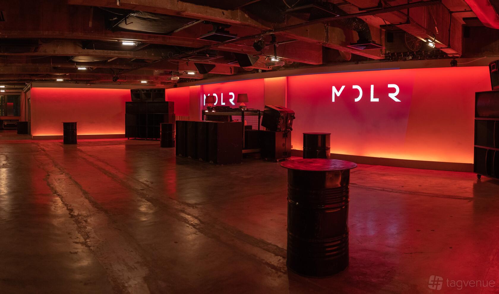 A nightclub with red lighting, industrial barrels as tables, and a DJ booth in front of illuminated MDLR signage.