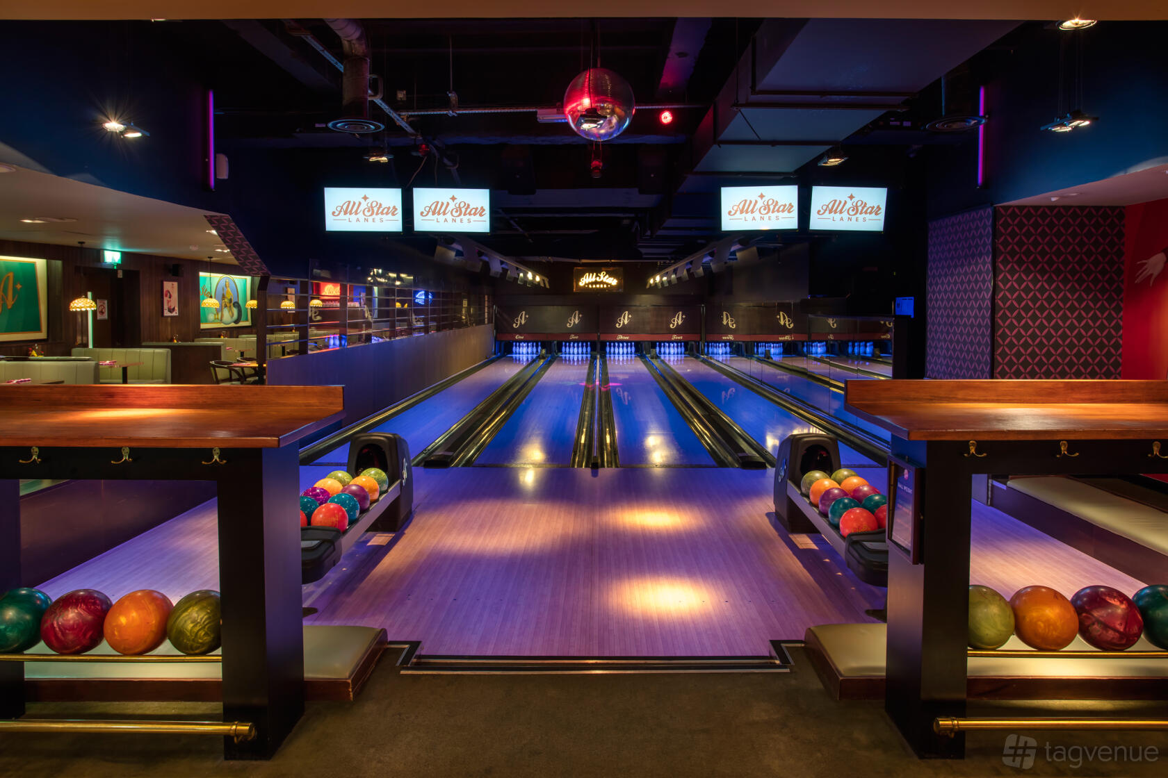 A bowling alley with colorful bowling balls, sleek wooden lanes, and overhead screens at All Star Lanes Holborn.