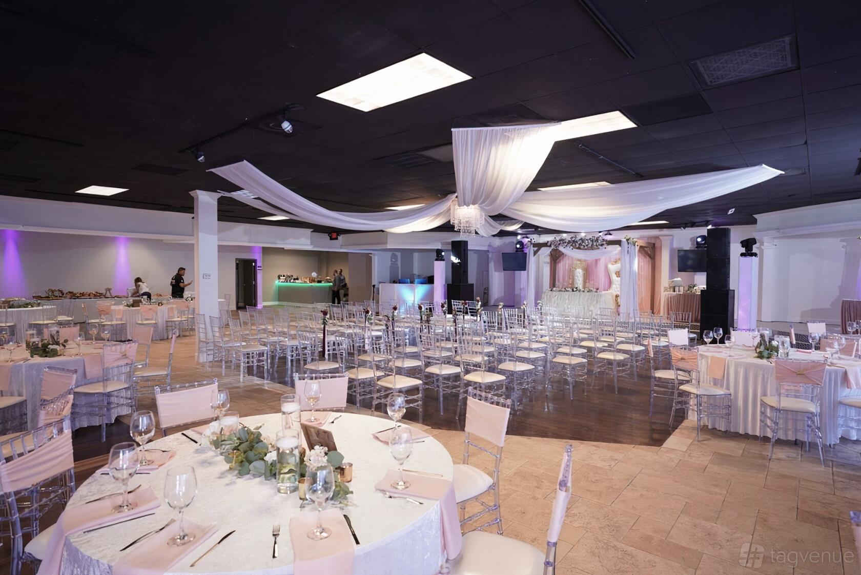 A spacious hall with round tables draped in light linens, clear chairs, and sheer white ceiling drapes at Reyes Event Hall.