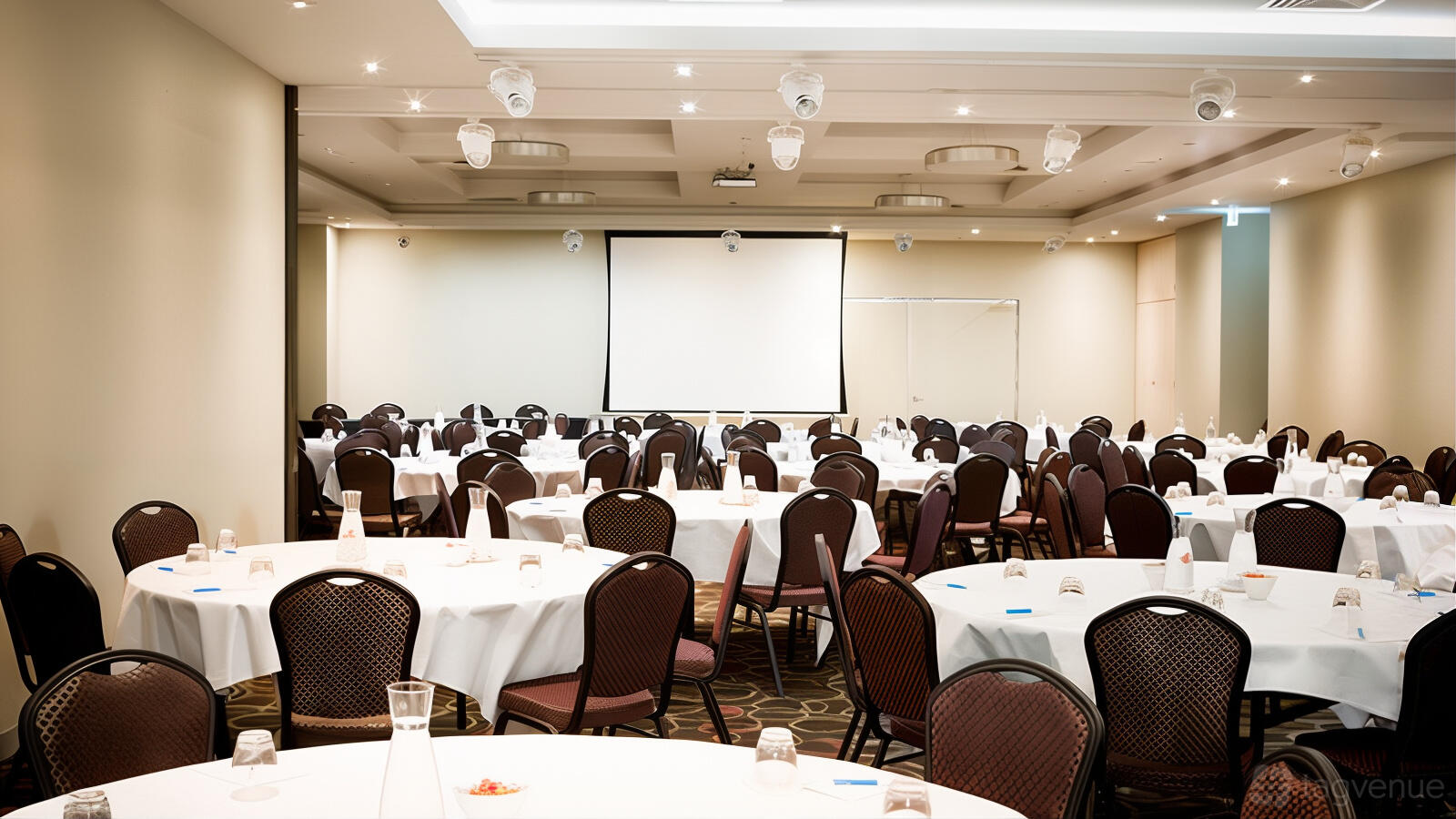 An event space with round tables covered in white linens, water pitchers, and a projector screen at Mantra Melbourne Airport.