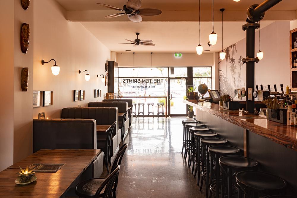 A bar with a long wooden countertop, barstools, booth seating, pendant lights, and wall art at The Ten Saints.