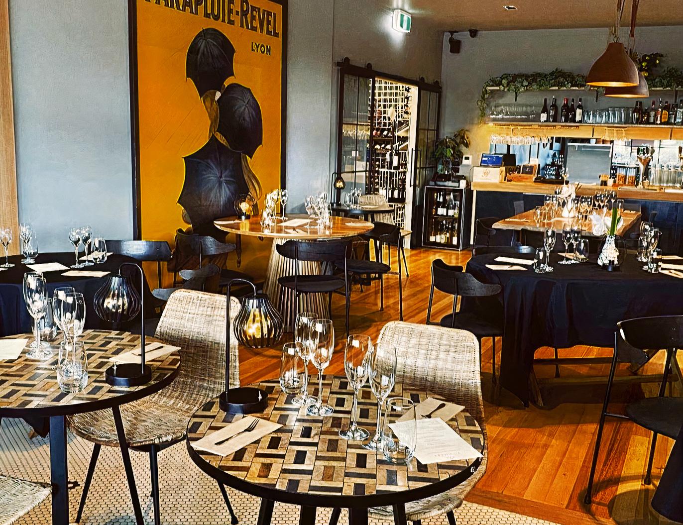 A bar with wooden floors, a mix of square and round tables set with glassware, and a stocked bar at Primrose & Vine.