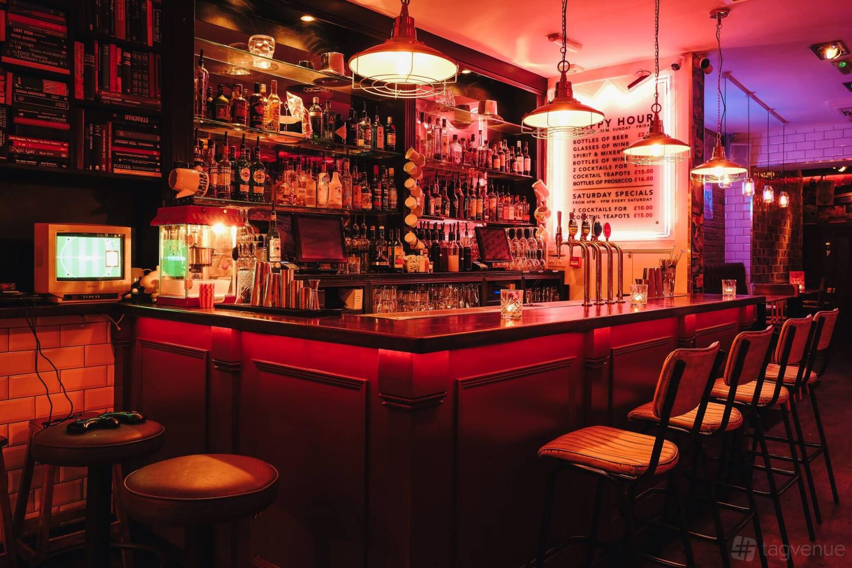 A cocktail bar with a backlit bar counter, exposed shelving of spirits, bar stools, and pendant lighting at Simmons | Angel.