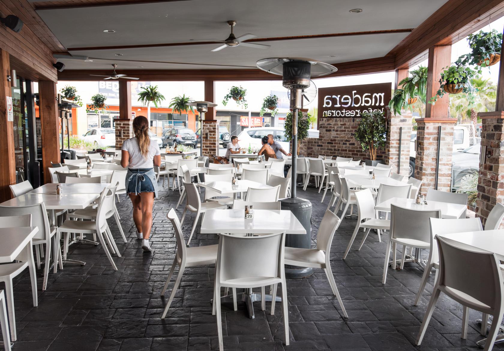 A dining room with white chairs, square tables, brick columns, and hanging plants at Madera Lounge.