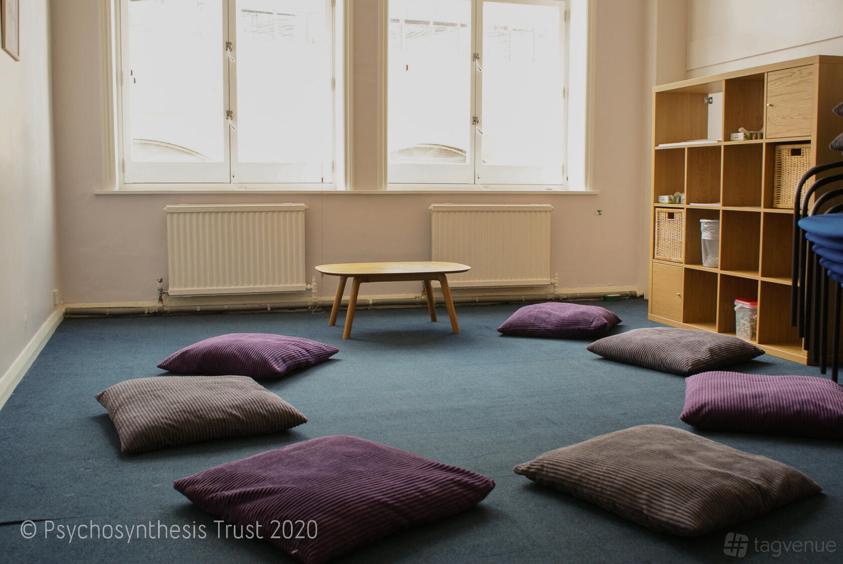 A training room with blue carpet, purple floor cushions in a circle, natural light, and a wooden shelf at The Psychosynthesis Trust.
