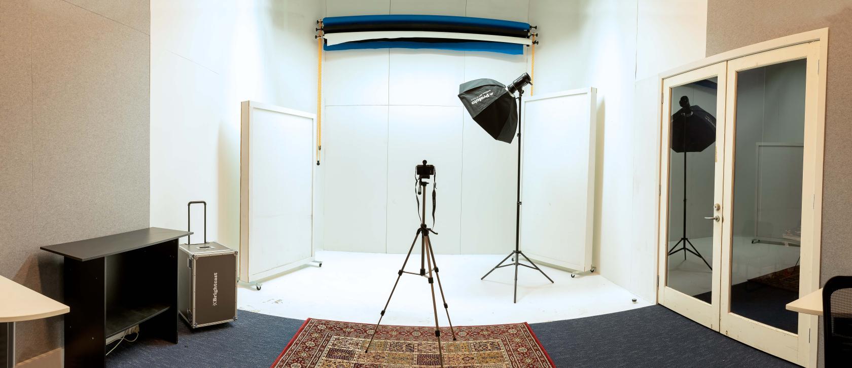 A photography studio with backdrop rolls, a camera on tripod, umbrella lighting, and a patterned rug at Empire Music Studios A.K.A Studio52.