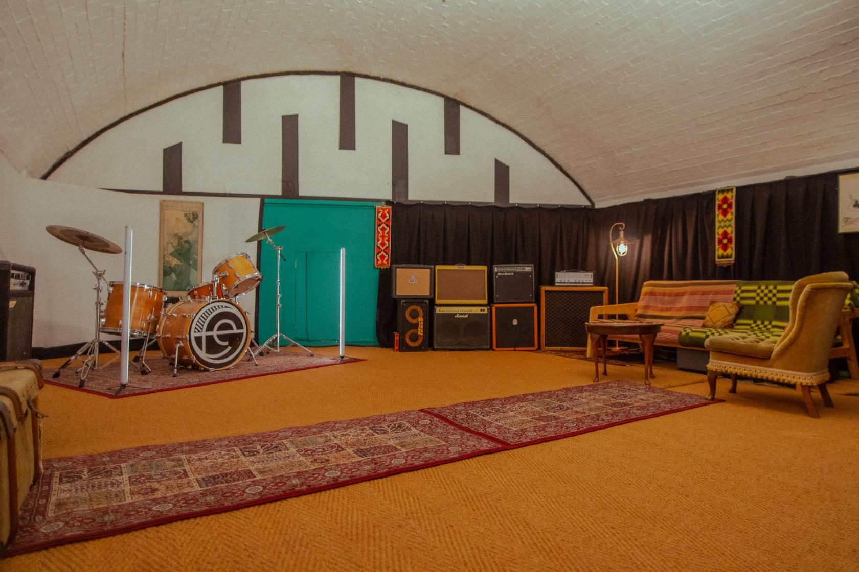 A recording studio with a drum set, vintage amplifiers, patterned rugs, and retro sofas at Arcus Sounds - Live Music Studio East London.