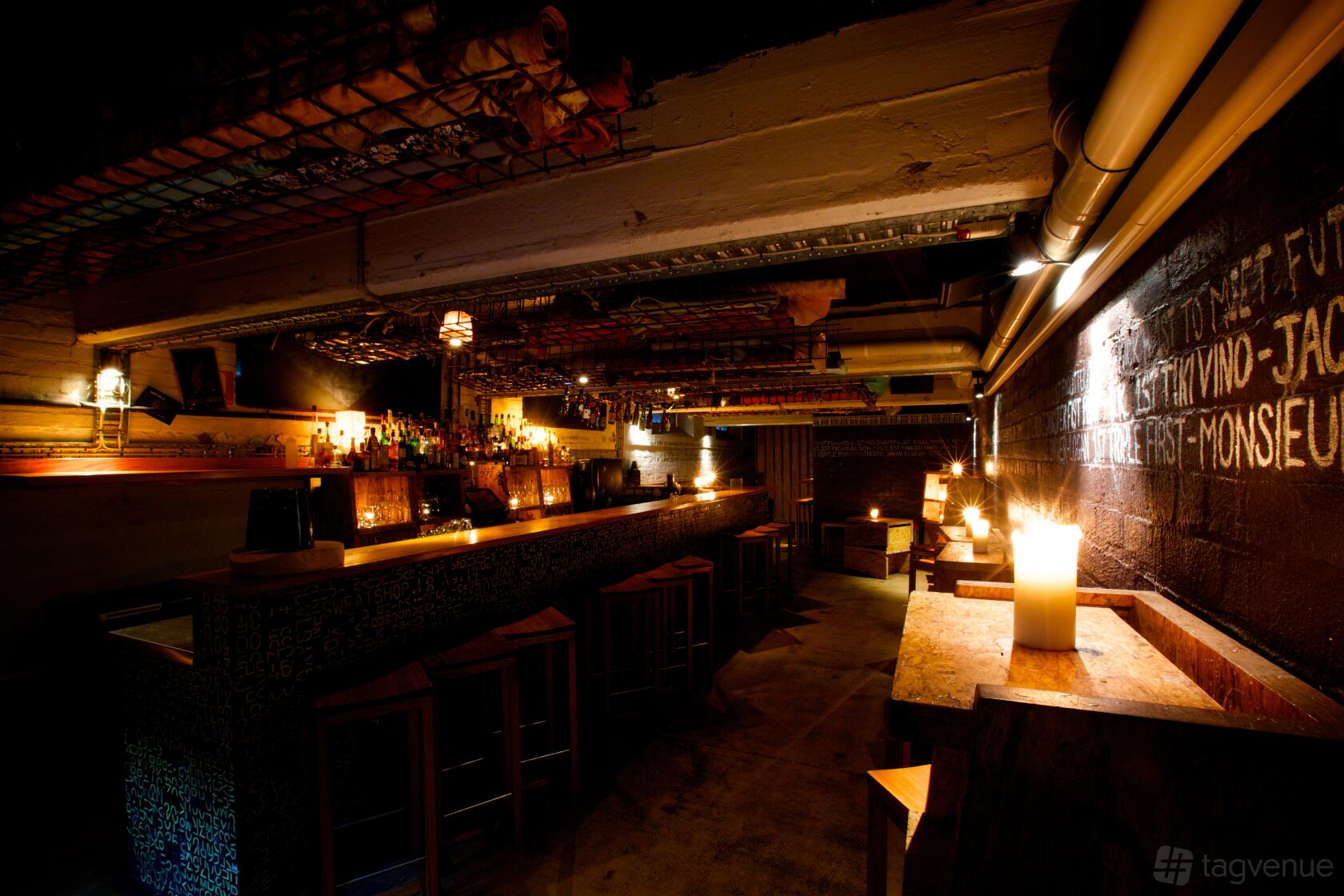 An underground bar with a long counter, high wooden stools, exposed pipes, and candlelit tables at Seamstress Restaurant & Bar.