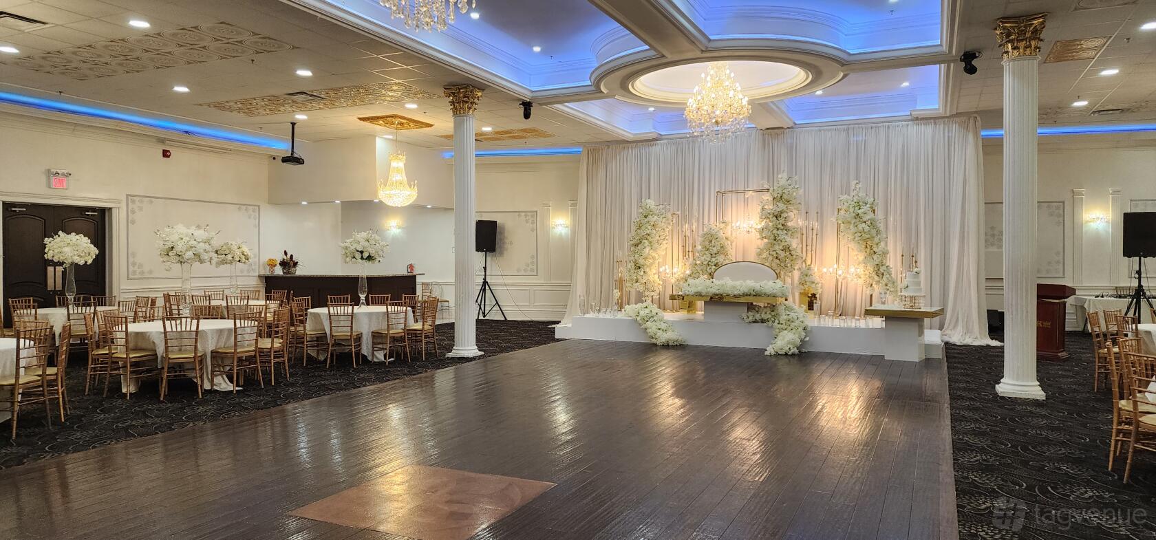 An event venue with a dark wood dance floor, chandeliers, and a white floral stage backdrop at Royal King Event Centre.
