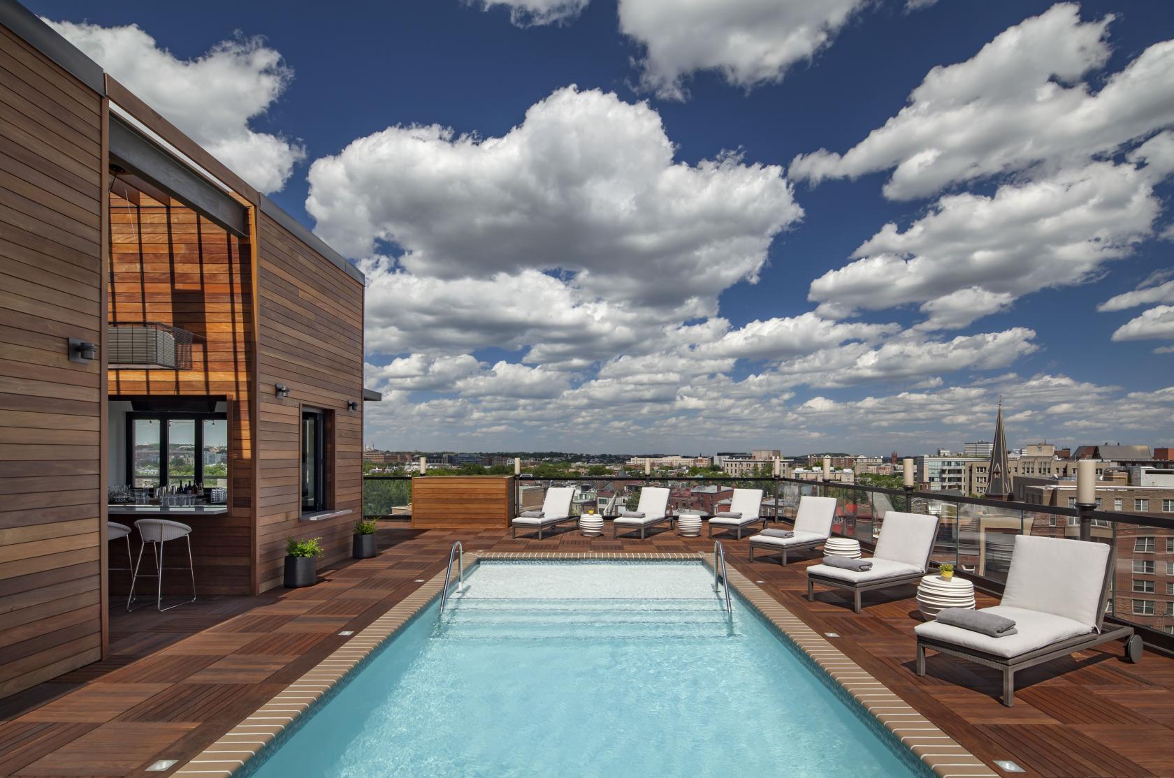 A rooftop bar with a pool, wooden deck, lounge chairs, and city skyline views at Hush Rooftop Bar.