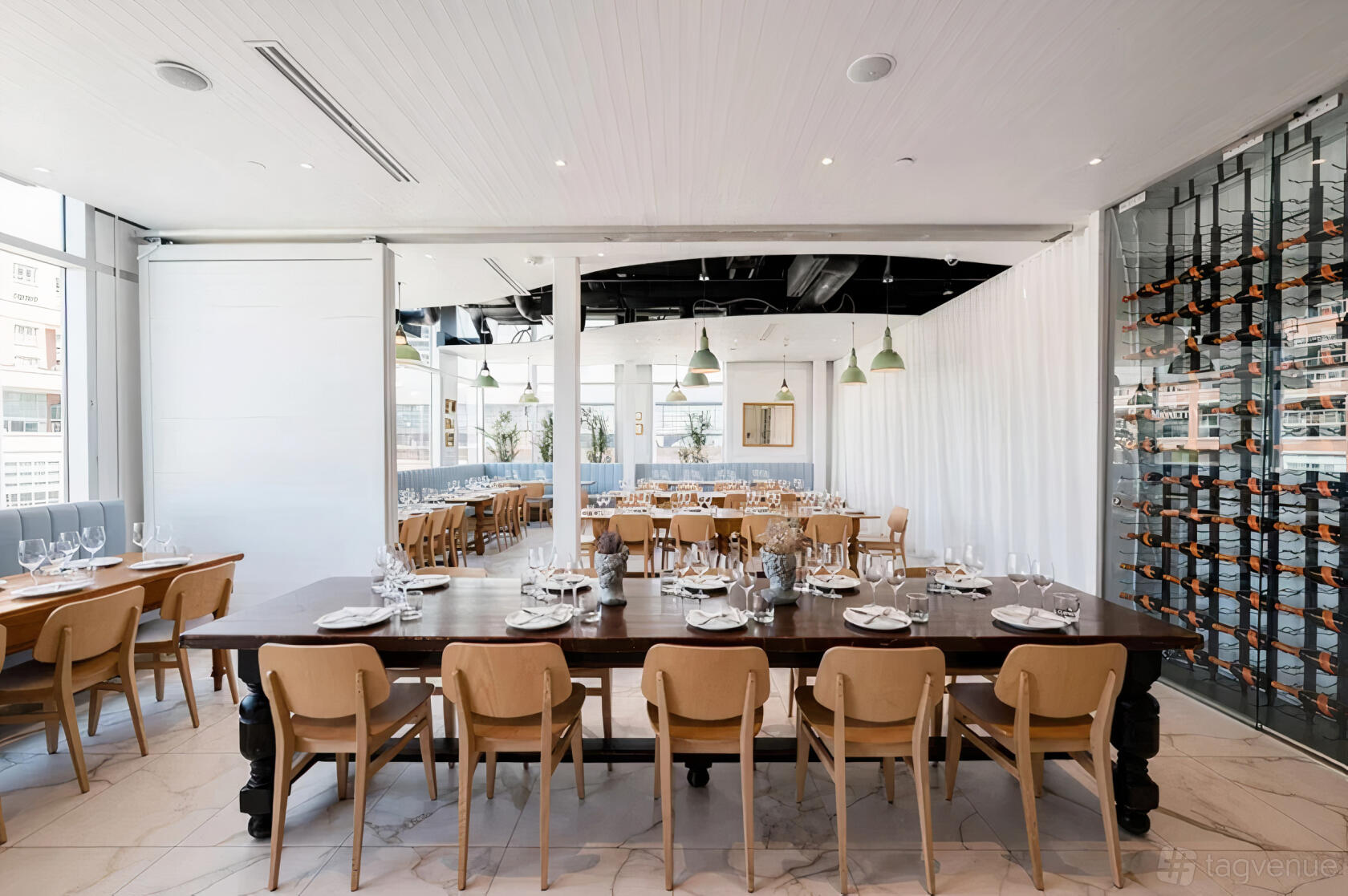A dining room with long wooden tables, tan chairs, floor-to-ceiling wine display, and white curtains at Nodo Liberty.