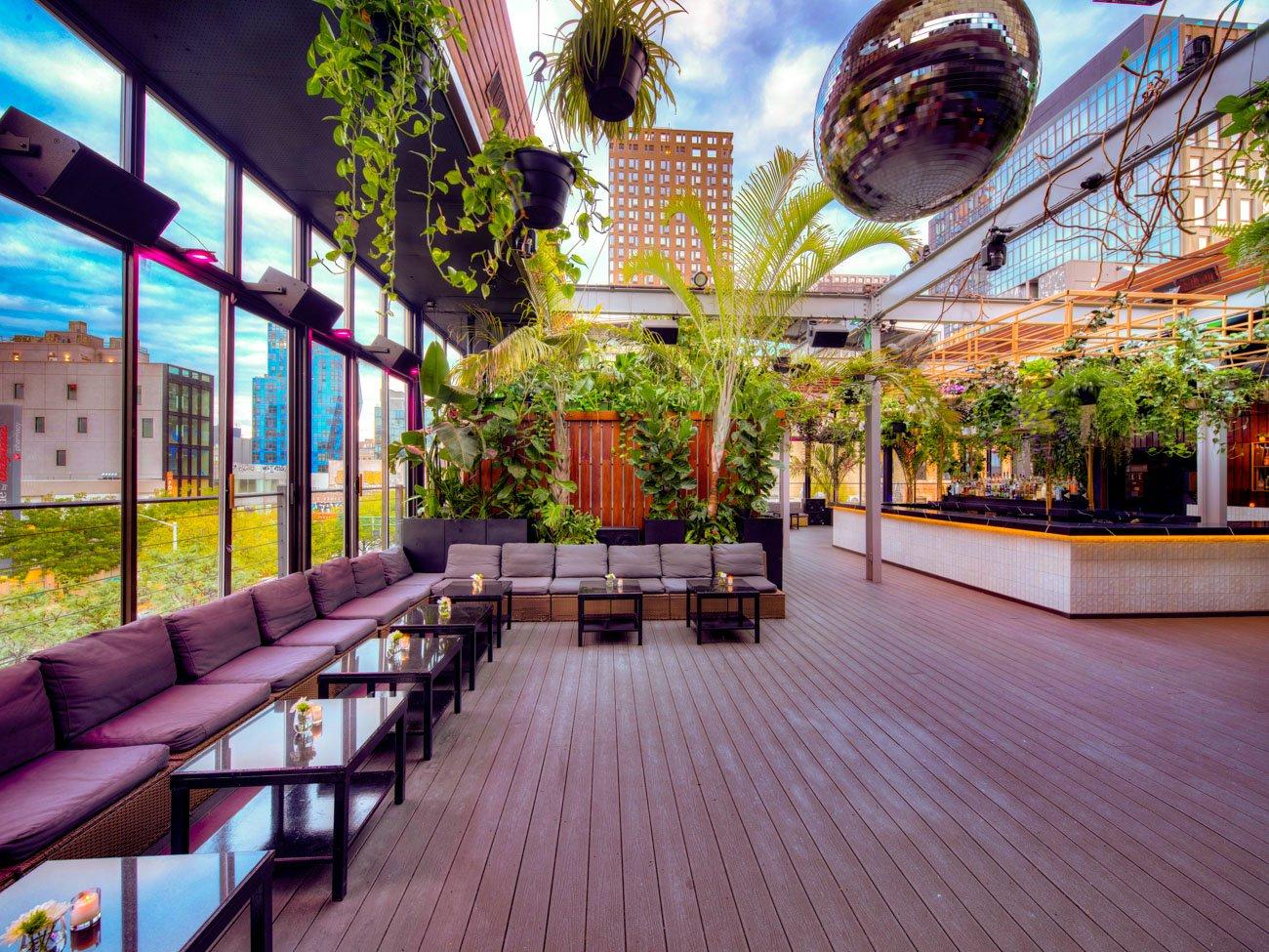 A rooftop lounge with potted plants, hanging greenery, cushioned seating, and a disco ball at The DL Rooftop.