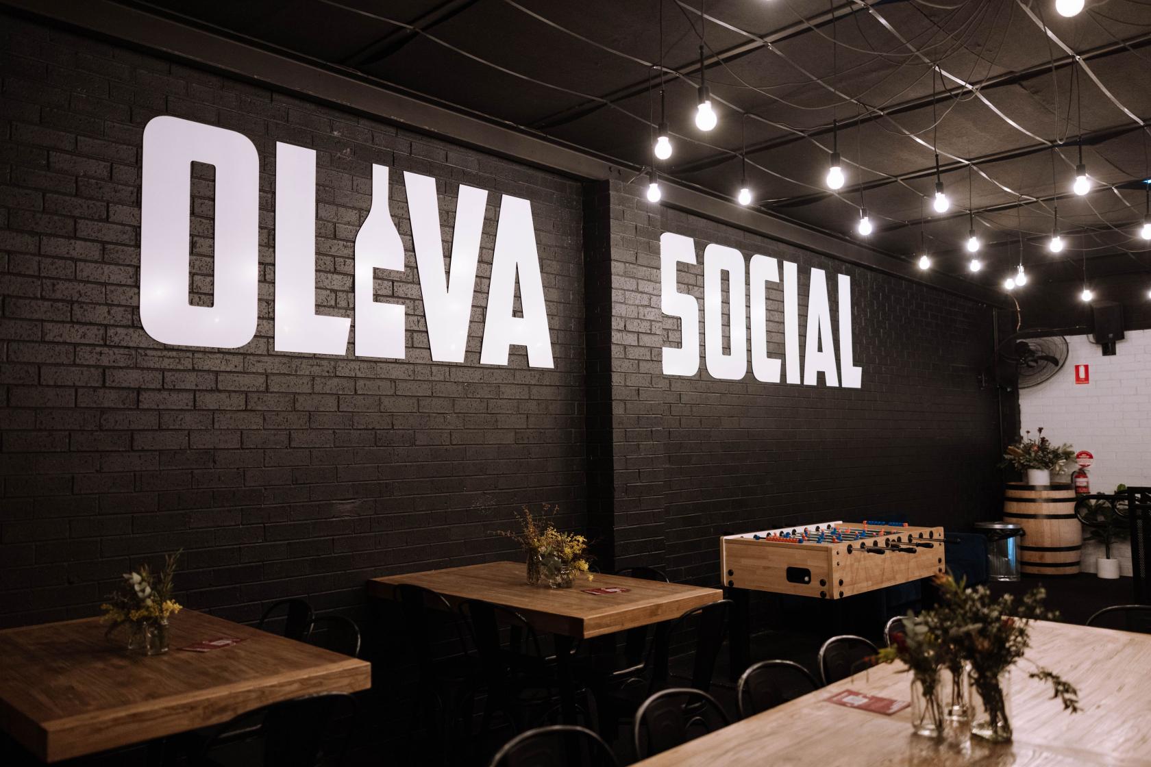 An event venue with wooden tables, black chairs, string lights, and a foosball table at Oliva Social.
