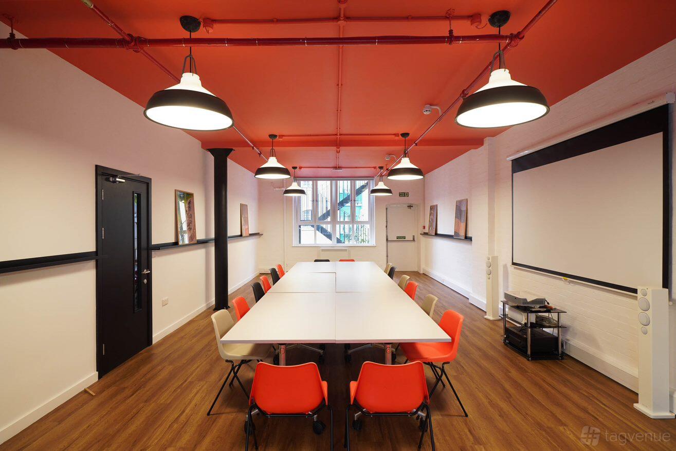 An event space with long white tables, orange chairs, wood flooring, and orange ceiling lights at London Bridge Hive.