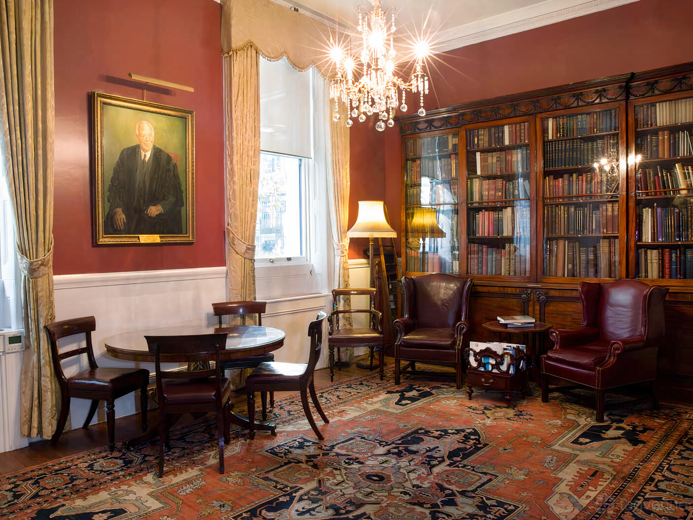 A library with antique bookshelves, leather armchairs, a chandelier, and a large portrait at Armourers' Hall.