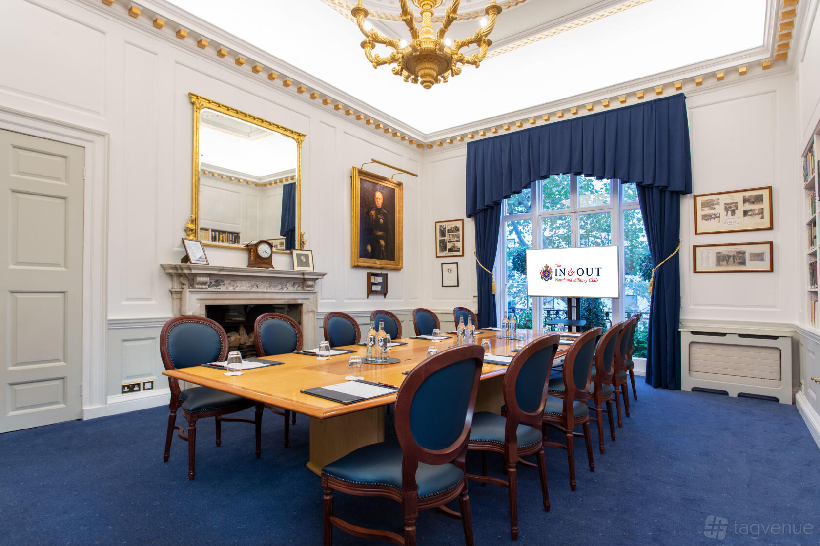 A traditional meeting room with a long wooden table, blue chairs, fireplace, and chandelier at In and Out (Naval & Military) Club.