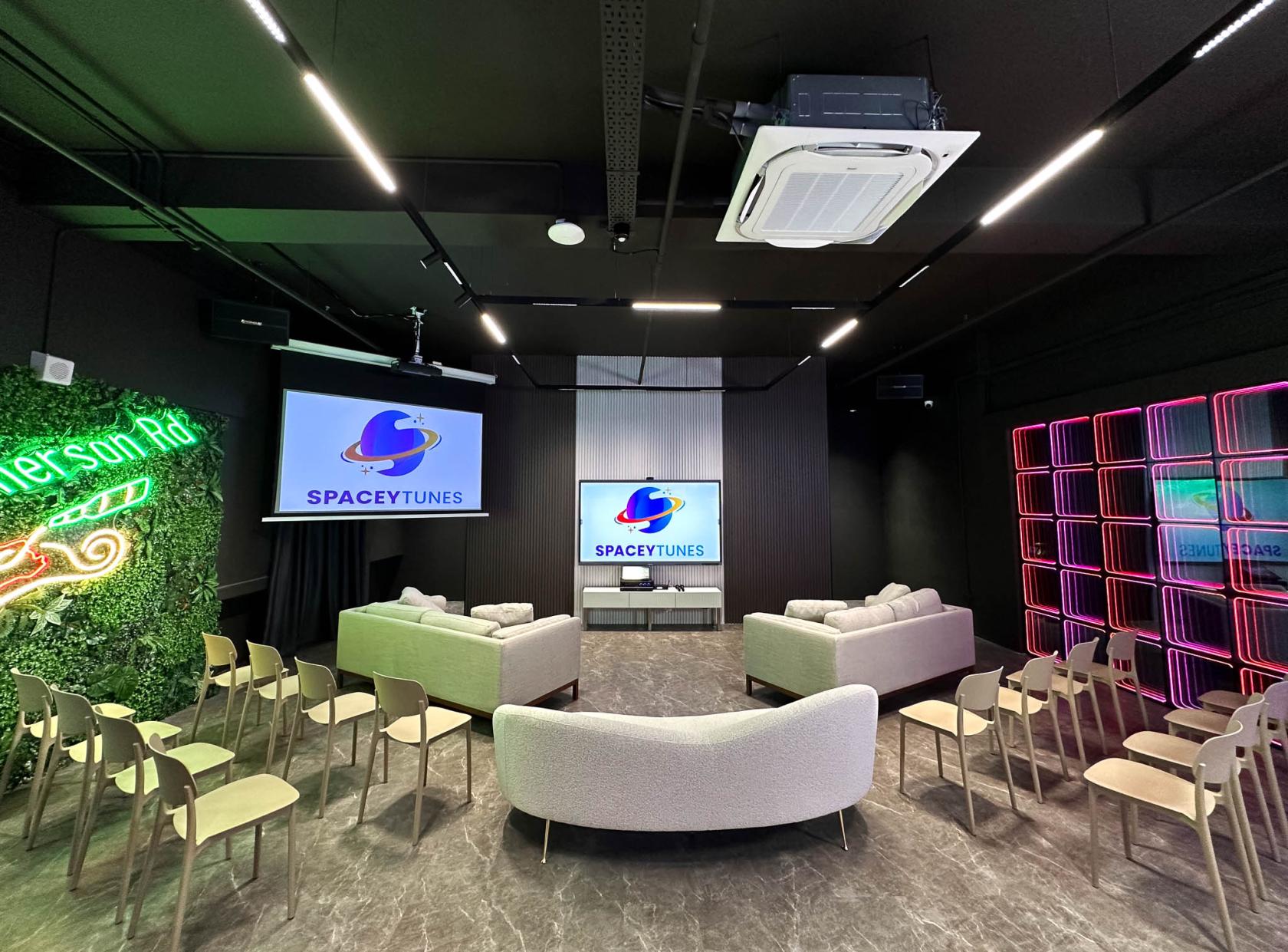 An event venue with curved sofas, rows of chairs, neon wall accents, and dual screens at Spaceytunes.