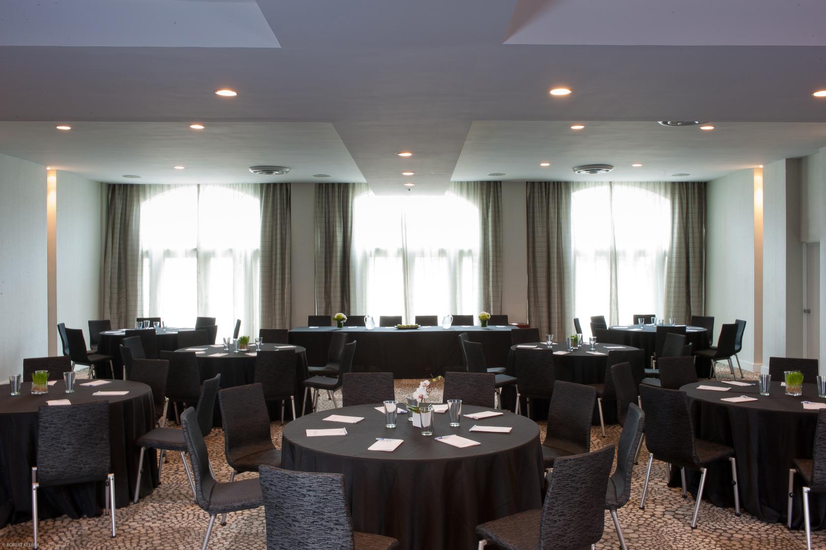 A hotel ballroom with floor-to-ceiling windows, round tables with black linens, and conference chairs at The Marseilles Hotel.