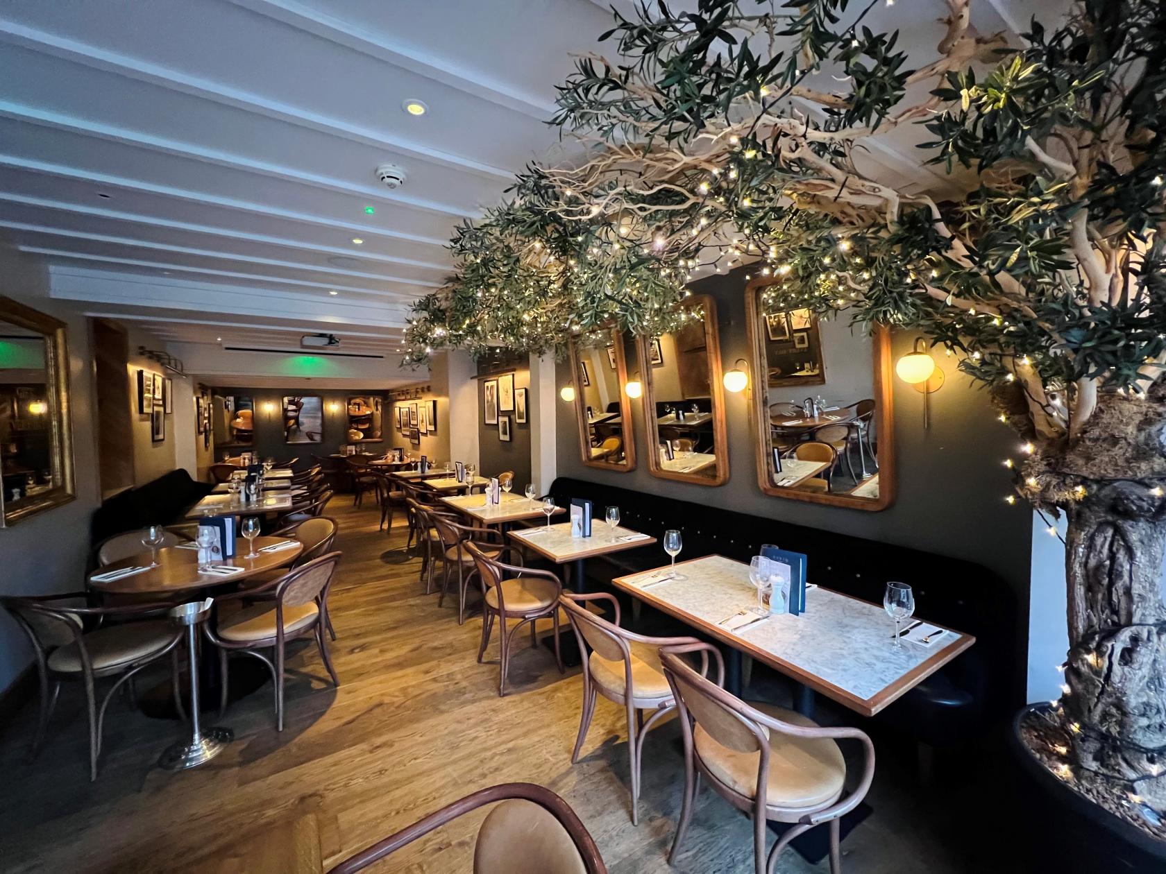 A restaurant with marble-topped tables, wooden chairs, and indoor trees with string lights at Gusto Italian, Birmingham.