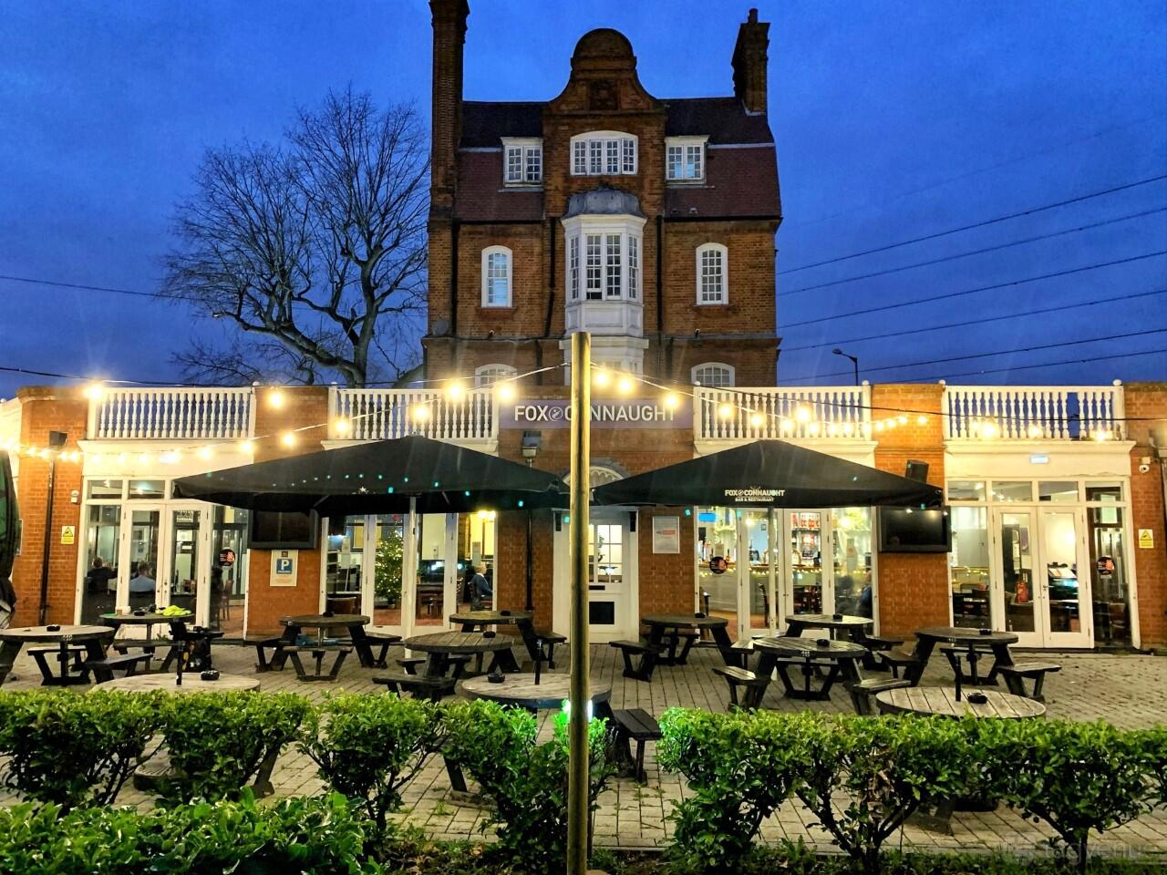 A pub terrace with black picnic tables, large umbrellas, string lights, and trimmed hedges at Fox Connaught.