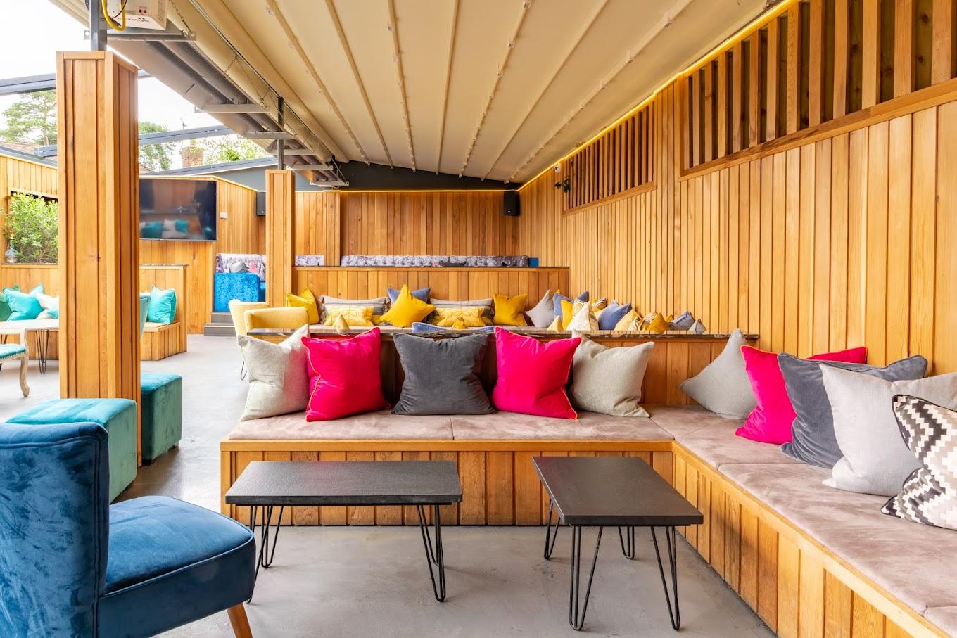 An outdoor bar with wooden paneling, cushioned bench seating, and colorful throw pillows at Fire & Ice Lounge Bar.