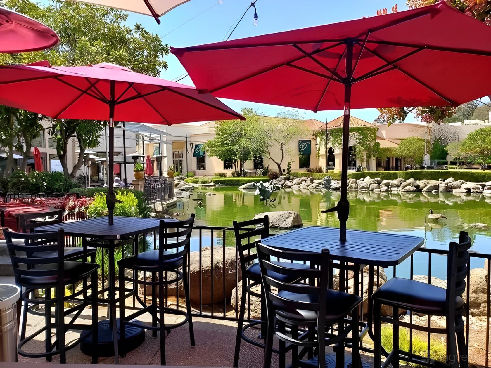 An outdoor restaurant patio with black tables, chairs, and red umbrellas overlooking a pond at 786 Kabob House.