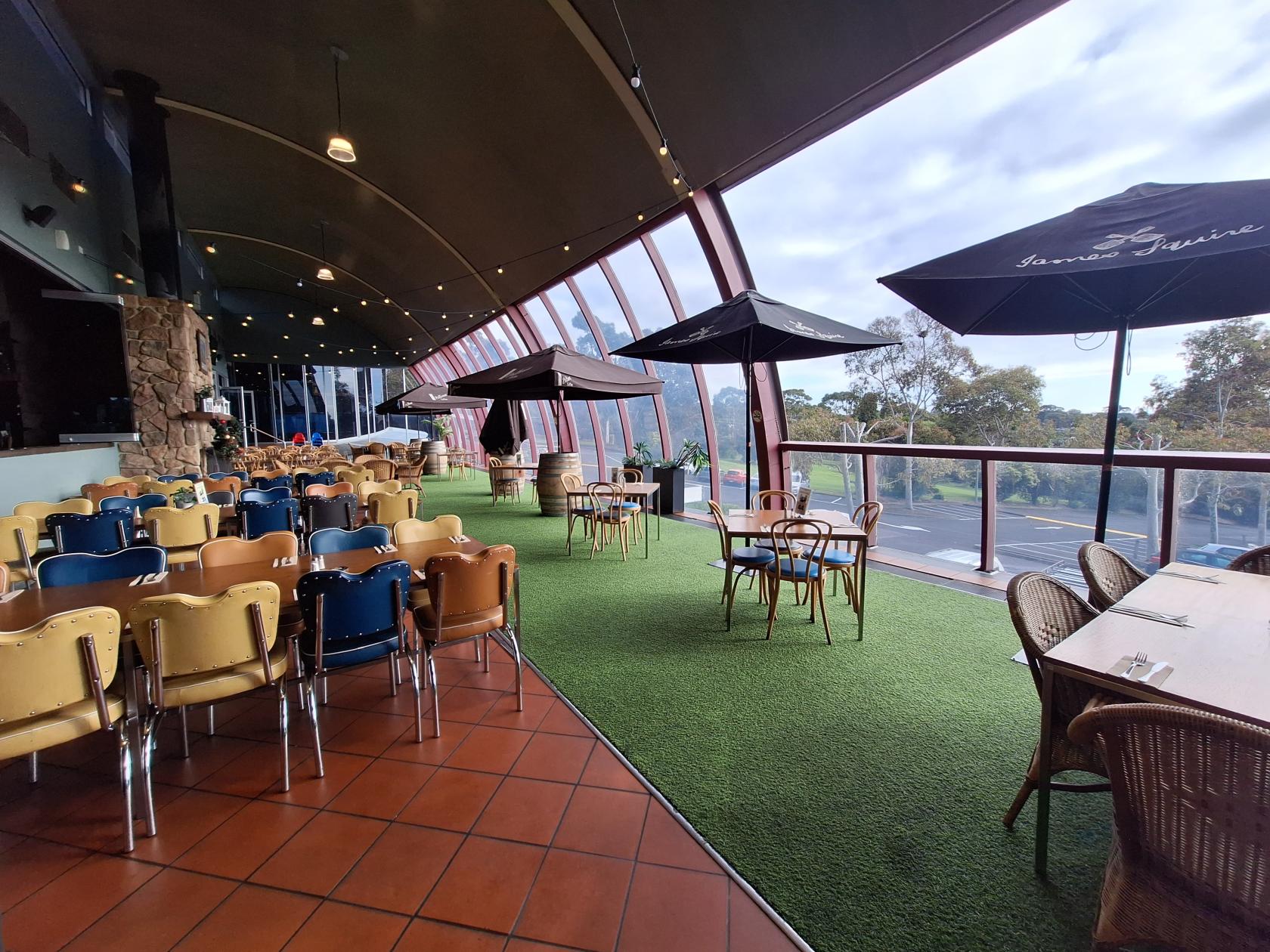 A pub terrace with artificial grass flooring, outdoor tables, colored chairs, and large umbrellas at Wheelers Hill Hotel.