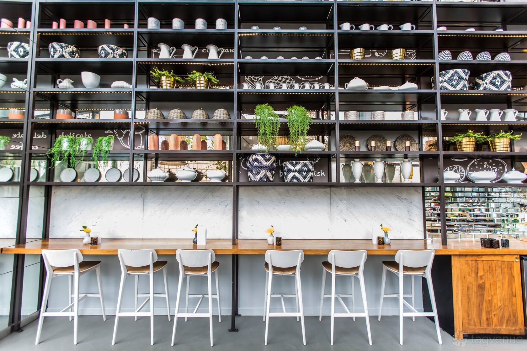A cocktail bar with a long wooden counter, high white chairs, and wall-to-ceiling open shelves of ceramics and plants at The Drift.