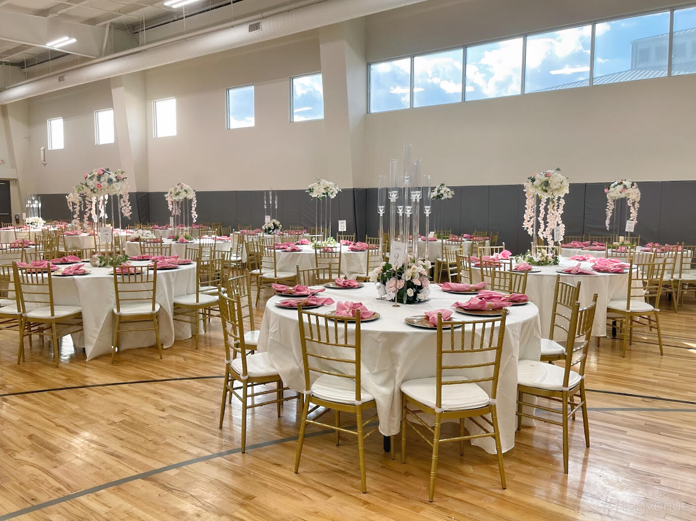 An event venue with round tables dressed in white linens, gold chairs, and pink floral centerpieces at TKP Pavilion Events and Sports Center.