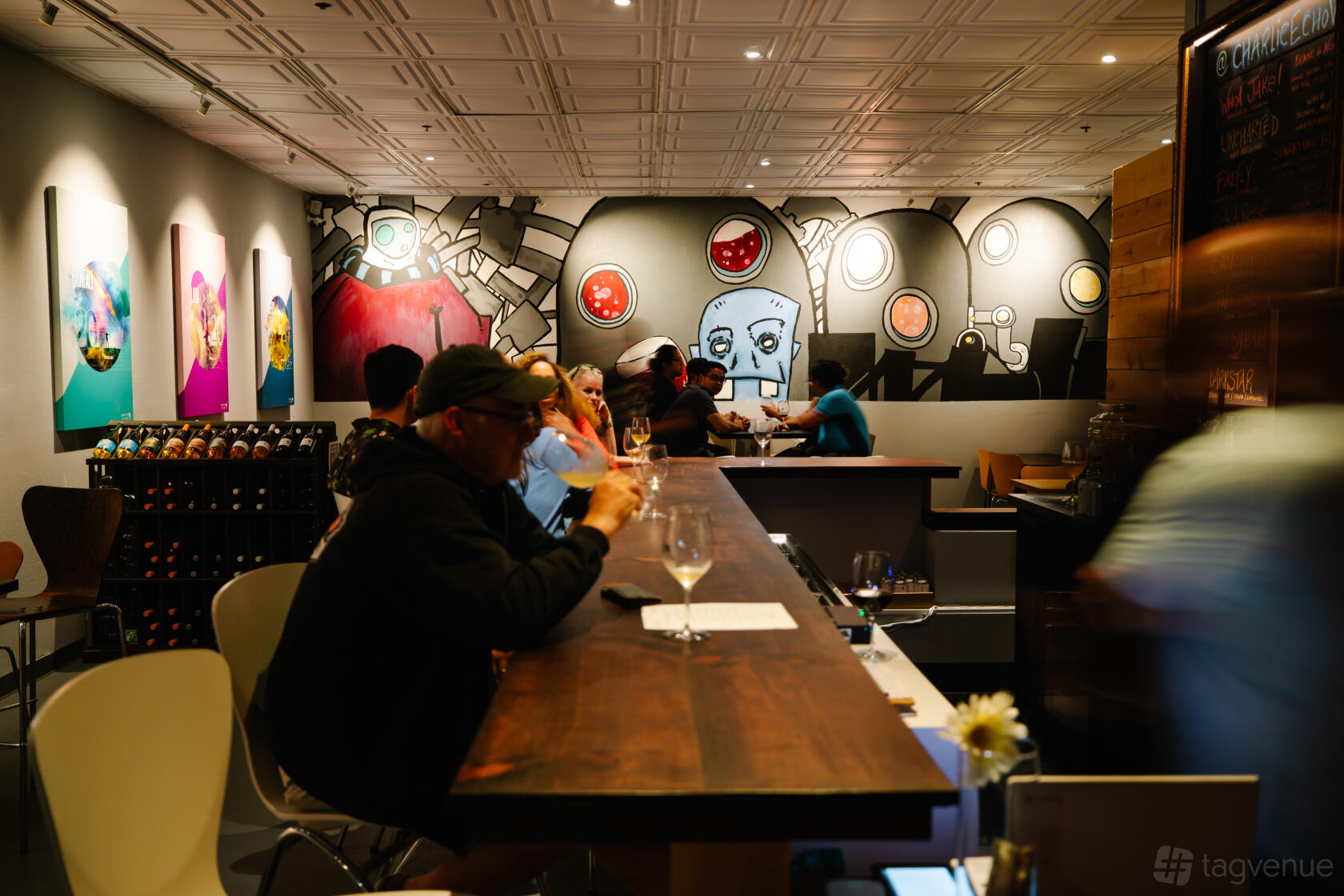 A bar with a wooden counter, patrons seated, colorful wall murals, and wine bottles on display at Charlie & Echo.