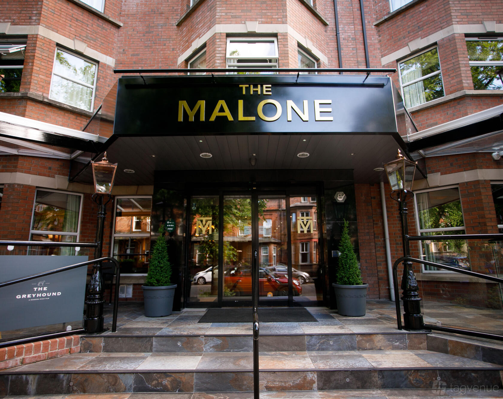 A hotel entrance with glass double doors, black canopy signage, and potted plants at The Malone Hotel.